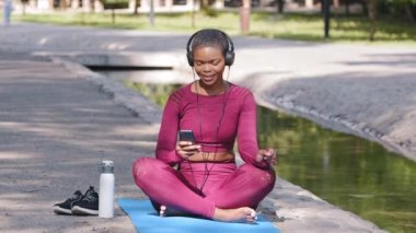 Pozitif büyük beden spor giyimli kadın portresi açık havada yoga minderinde rahatlıyor, sadece kulaklıkla müzik dinleyen güzel bir bayan fitness eğitmeni gülümsüyor. Spor aktif yaşam tarzı konsepti