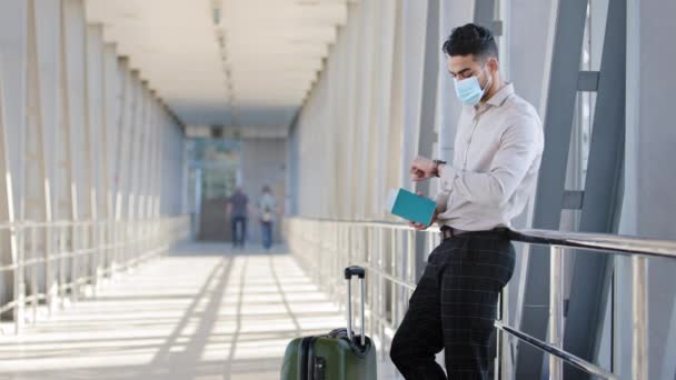 Hispanic male passenger business man wears formal clothes and face protective medical mask holds passport boarding pass plane ticket stands with suitcase in airport terminal waiting for flight travel — Stock Video