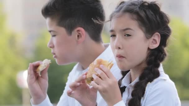 Двое голодных детей школьники школьники студенты брат и сестра латиноамериканцы дети друзья сидят на открытом воздухе в школьном дворе едят пироги булочки пирожные наслаждаясь едой пить чистую воду из бутылки — стоковое видео