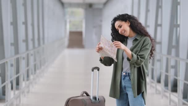 Молодая кавказка девушка женщина пассажирка туристка с чемоданом стоит у терминала аэропорта железнодорожный вокзал смотрит на бумажную карту поиска маршрута дороги направления отель думает навигация — стоковое видео