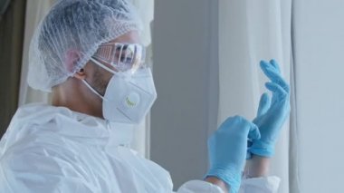 Erkek doktor, genç tıp çalışanı özel koruma kıyafeti giyiyor, üniforma, gözlük takıyor, ellerine lastik mavi eldiven takıyor, covid-19 koronavirüs salgınıyla çalışmaya hazırlanıyor.