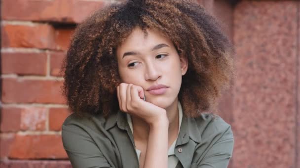 Одягнена задумлива молода афроамериканська жінка проводить вільний час на самоті, сидячи надворі, втративши глибокі думки, відчуваючи нудьгу. Дивлячись на пригнічені думки про особисті проблеми, меланхолійна концепція настрою — стокове відео