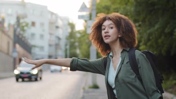 Schöne afroamerikanische Millennial-Frau in Freizeitkleidung winkt Auto herunter, trampt am Straßenrand. Hübsche junge Frau mit schwarzem Lockenkopf, die allein unterwegs ist und Sommerurlaub hat. Erlebnis- und Tourismuskonzept — Stockvideo