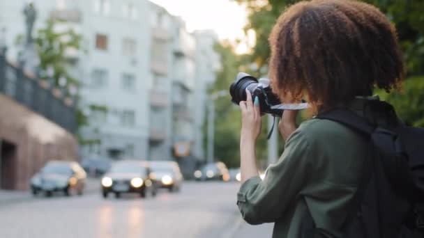 Joven turista africana con pelo rizado y mochila viajando, fotografiando atracciones usando dispositivo digital. Blogger de viajes tomando fotos con cámara fotográfica profesional en el centro de la ciudad de verano — Vídeos de Stock