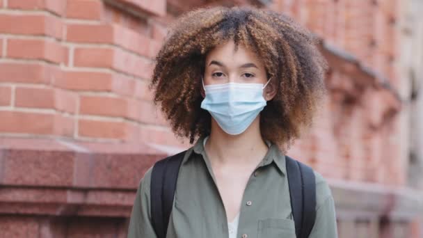 Kopfschuss-Porträt afroamerikanische junge Frau in blauer medizinischer Schutzmaske, die vor Backsteinbau-Hintergrund steht, blickt in die Kamera. Coronavirus-Ausbruch stoppen, Konzept zur Luftreinhaltung — Stockvideo