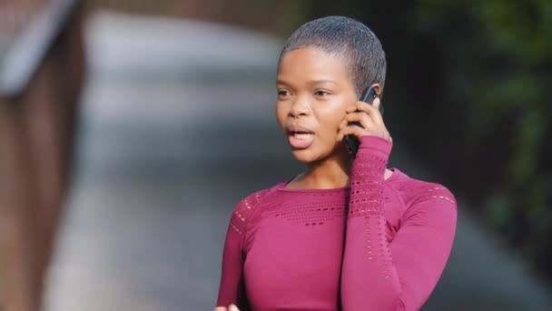 Snímek hlavy portrét 25s Africké černošky dívka ve sportovním oblečení stojící venku mluvit na smartphone během ranního cvičení v parku. Mladá žena běžkyně má příjemnou konverzaci po telefonu, směje se, cítí šťastný — Stock video
