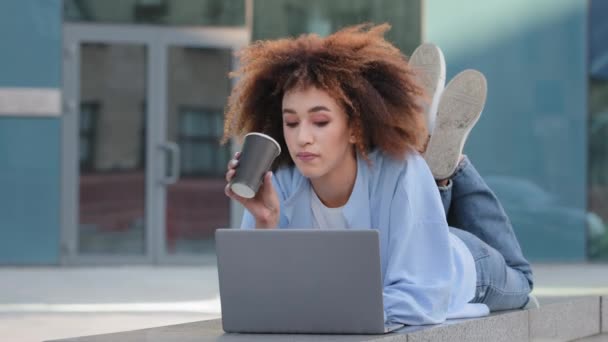 Afrikansk flicka student afro amerikansk affärskvinna med lockigt hår frilansare användaren ligger utomhus nära företagets kontor byggnad skriva på bärbar dator arbetar med dator online chatta dricka kaffe te — Stockvideo