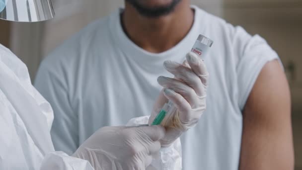 Gros plan infirmière médecin en uniforme de protection médicale gants en latex tenant bouteille dose de vaccin de covid19 remèdes contre le coronavirus mettre du liquide dans la seringue pour injection à l'homme souriant africain — Video