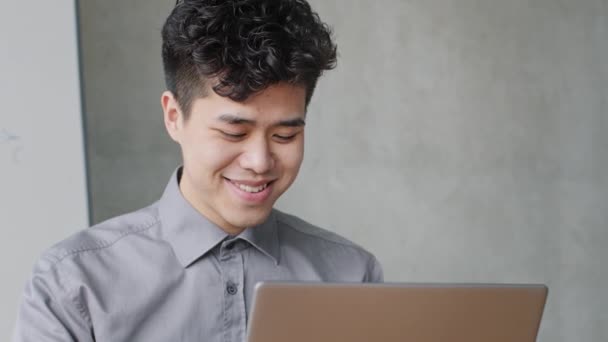 Retrato dentro de casa asiático cara estudante jovem coreano homem de negócios chinês japonês gerente escritório trabalhador chefe líder empresário usa laptop para o trabalho olha para tela do computador lê engraçado notícias online sorriso — Vídeo de Stock