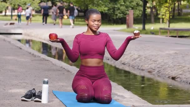 Vonzó fiatal felnőtt nő fitness oktató ül jóga szőnyegen a nyári parkban tartja alma és sütemény, cupcake, próbál a helyes választás. Millenniumi lány választja az egészséges életmód, egészséges ételek — Stock videók