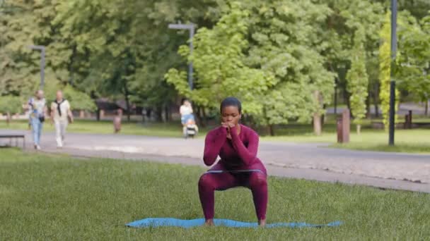 Atlético menina negra africana em sportswear exercício, executa agachamentos com elástico. Atraente focado biracial jovem mulher fazendo alongamento durante o treino matinal no parque. Pilates, ioga, fitness — Vídeo de Stock