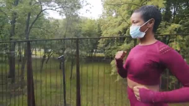 Mujer hermosa atlética negra en ropa deportiva y máscara médica protectora comprometida velocidad caminando en el puente al aire libre durante el entrenamiento diario por la mañana. Joven fitness femenina cuida de su cuerpo, se calienta — Vídeos de Stock