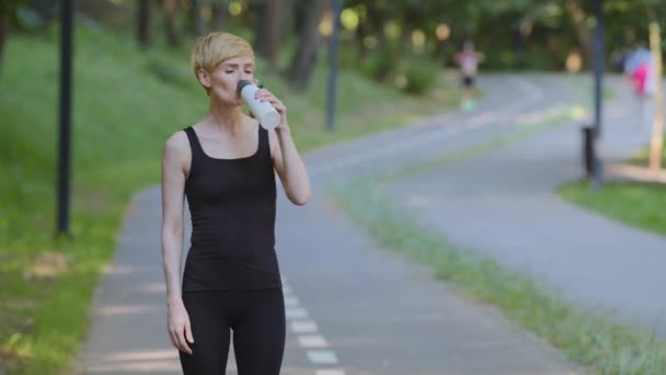 Femme caucasienne d'âge moyen sportive athlète coureuse joggeuse se tient dans le parc de la ville se sentant fatiguée après le jogging courir séance d'entraînement en plein air boissons eau froide encore essuie la sueur du front après le sport — Video