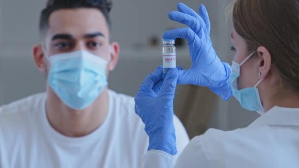 Médecin méconnaissable femme infirmière en masque médical et gants en latex tient montre bouteille avec covide-19 vaccin coronavirus remède médicament pour arabe espagnol espagnols homme malade patient assis à la table à la clinique — Video