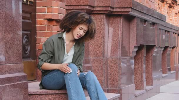 African American woman holding and massaging her leg because of injury. Young black girl having knee trauma during walking around city. Illness, painful body condition, ache, joint ligament problem — Stock Video