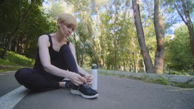 Orta yaşlı beyaz kadın atlet parkta oturmuş spor ayakkabılarına bağcıklar bağlamış açık havada spor yaptıktan sonra şişeden soğuk su içmiş.