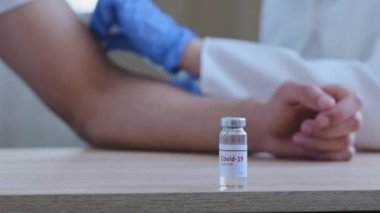 Coronavirus 'a karşı ilaç içeren bir şişe covid-19 aşısı hastane kliniğinin arka planında bulanık erkek hasta ve doktor aşısının ahşap masada duruyor.