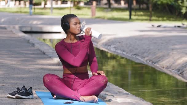 Atletisch plus size Afro-Amerikaanse mooie vrouw afwerking van de training en het drinken van eiwit melk schudden vitaminen of water na de training, het luisteren naar muziek met oortelefoons. Sport actief lifestyle concept — Stockvideo