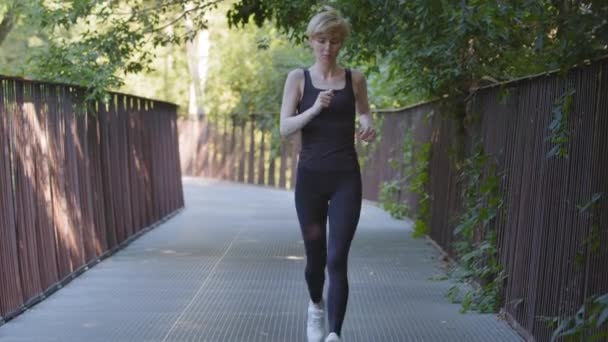 Középkorú atlétikai vékony nő edző kaukázusi hölgy visel fekete melegítő fut a hídon a parkban csinál sport kardio edzés kocogás gyors futás kocogás fáradtság megáll a légzést erősen — Stock videók