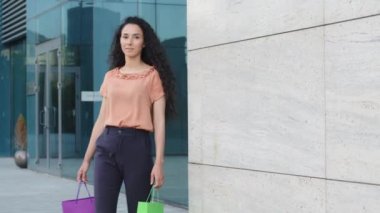 Güzel modaya uygun genç İspanyol bayan tüketici alıcı mutlu kadın alışveriş merkezinden parlak alışveriş çantalarıyla çıkıyor indirimlerden keyif alıyor yavaş çekimde hediye paketleri taşıyor.