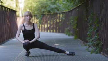 İnce, orta yaşlı, beyaz kadın antrenör kadın koşucu siyah spor giysisi giyiyor. Bir bacağının üzerinde oturuyor. Kalça kaslarını kasıyor, parkta koşmadan önce esniyor.