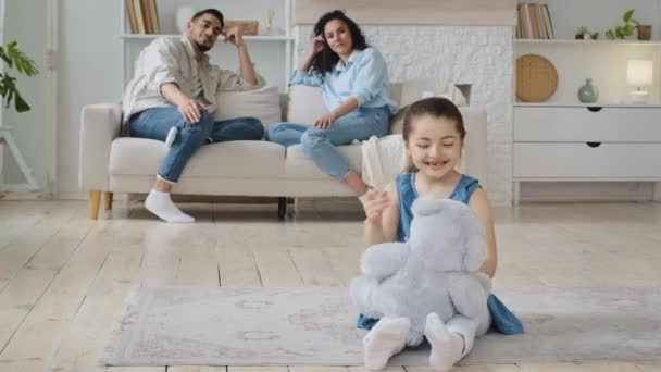 Kafkas asıllı ispanyol küçük kız çocuğu oturma odasında oturmuş oyuncak ayıyla oynuyor genç çok ırklı ebeveynler rahat koltukta dinleniyor çocukluk zamanlarının tadını çıkarıyor. — Stok video