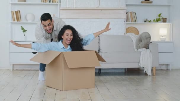 Wanita berambut keriting lucu duduk di dalam kotak karton. Pria Hispanik mendorong bingkisan dengan wanita. Pasangan bahagia menyewa pembeli memiliki menyenangkan packing relokasi pada hari pindah di apartemen baru, relokasi hipotek — Stok Video