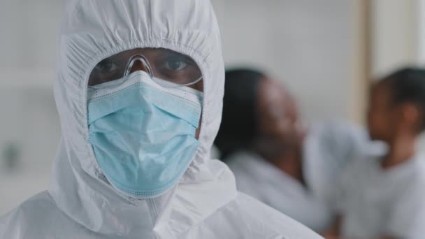 Afro american doctor scientist medical worker wears protective clothing hood transparent glasses mask looks at camera stands posing on blurred background woman with baby african mother with daughter — Stock Video