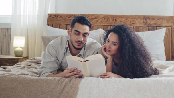 Español pareja casada barbudo marido y rizado esposa novio y novia hispano hombre y mujer se acuestan en la cama en casa en el dormitorio leer interesante libro literatura disfrutar leyendo juntos pasar tiempo — Vídeos de Stock