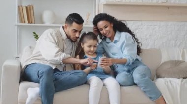 İspanyol aile kanepede oturup fotoğraf çekmek ve video kaydetmek için telefon kullanıyor. Küçük kızı olan ebeveynler komik maskeler denerken online mobil uygulama gülen çocuk dil gösteriyor.