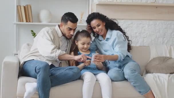 Hiszpańska rodzina siedzi na kanapie, korzystając z telefonu do robienia zdjęć i nagrywania wideo. Rodzice z małą córeczką bawią się próbując śmieszne maski online aplikacja mobilna śmiejąc dziecko pokazując język — Wideo stockowe