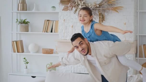Opiekuńczy Hiszpan mężczyzna kochający ojciec trzyma na plecach mała córka bawi się z dziewczyna dziecko piggyback udawać samolot mucha rozszerzając ramiona na boki bawiąc się w domu razem śmiejąc się wolny czas — Wideo stockowe