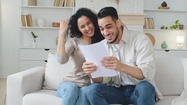 Eufórico español pareja étnica sentarse en el sofá en casa lectura documento carta de papel con noticias increíbles préstamo bancario aprobación rentable oferta de trabajo oportunidad celebrando el éxito abrazo hacer gesto de victoria — Vídeo de stock