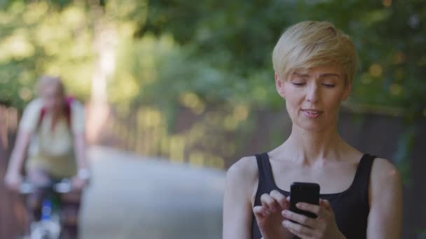 Close-up portrait middle-aged caucasian female lady blonde woman with short hair standing outdoors in park looking at screen of smartphone using mobile phone gadget for chatting browsing Internet app — Stock Video