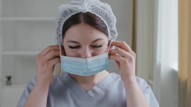 Молодая кавказка-медсестра, врач-интерн, медицинский работник в форме, позирует в больничной клинике, надевает защитную маску от инфекции коронавируса ковид-19, смотрит в камеру — стоковое видео