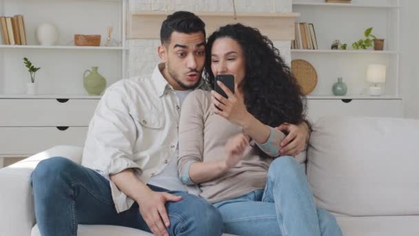 Счастливая молодая латиноамериканская супружеская пара, глядя на экран смартфона, сидящего на диване, получает сообщение с хорошими новостями онлайн лотереи раздачи смотреть смешное видео в сети с помощью мобильного телефона — стоковое видео