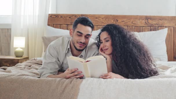 Многонациональная испанская арабская пара кавказский мужчина муж и женщина подружка жена лежат вместе на кровати в уютной спальне дома читая одну книгу насладиться новой литературой изучения отдыха в помещении — стоковое видео