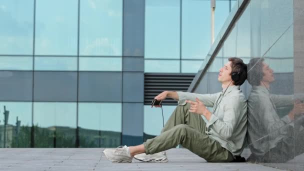 Heureux gars millénaire dans les écouteurs assis sur le sol en face de la construction en verre moderne profiter de la musique énergique rythmique. Jeune hipster écouter des morceaux préférés, profiter d'un son de qualité avec les yeux fermés — Video