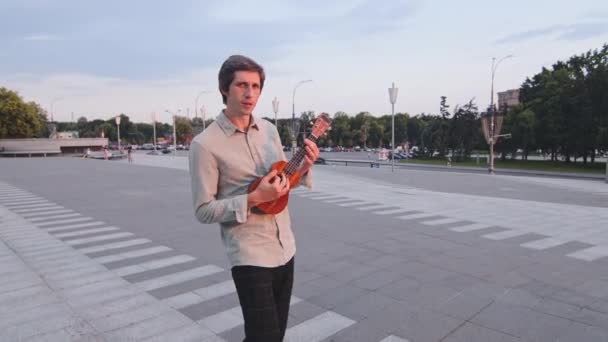 Feliz joven estudiante camina por las calles de la gran ciudad con el instrumento musical tradicional hawaiano. Músico masculino tocando la pequeña guitarra ukelele exótica, disfrutando de lo que ama, hobby, pasar el fin de semana solo — Vídeo de stock