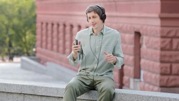 Homem milenar feliz vestindo fones de ouvido modernos se divertir ouvindo música faixas favoritas, relaxar ao ar livre, feliz jovem macho em fones de ouvido dança desfrutar de lazer fim de semana sozinho, conceito livre de estresse — Vídeo de Stock