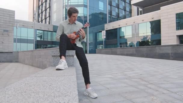 Young adult man performs with exotic musical instrument in downtown on modern glass office building background. Millennial musician wearing youth clothes and sneakers sitting outdoors playing ukulele — Stock Video