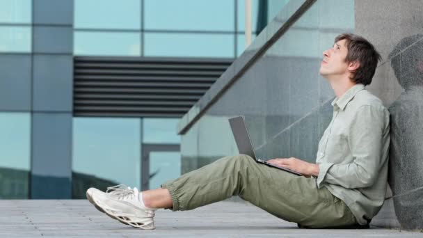 Joven periodista freelancer sentado al aire libre usando el ordenador portátil tecleando en el teclado del dispositivo electrónico mirando en el monitor. La lluvia inesperada distrae al chico del trabajo, le impide alcanzar el éxito — Vídeos de Stock