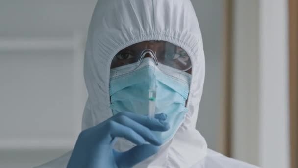 Medico chirurgo africano medico operaio in uniforme protettiva occhiali trasparenti e maschera indossa guanti di lattice tiene siringa con medicina rimedio liquido vaccino si prepara alla vaccinazione iniettabile — Video Stock