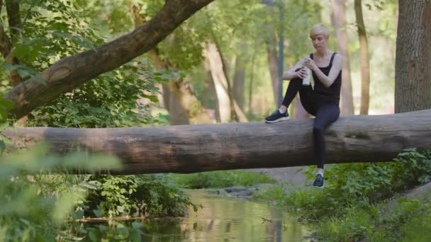 Snella sportiva donna sportiva caucasica seduta sul tronco d'albero nel parco all'aperto nella foresta verde natura a riposo dopo l'allenamento di jogging beve acqua fresca fredda dalla bottiglia sente la sete riposo rilassante — Video Stock