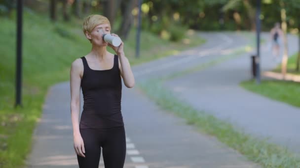 Slank atletisk træt kvinde sportskvinde runner jogger i sportstøj føles tørstig efter jogging kører træning står i parken udendørs drikkevarer koldt ferskvand drikker fra flaske forfriskning – Stock-video