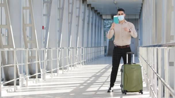 Успішний іспаномовний бізнесмен пасажирський інвестор носить медичну маску, показує паспорт посадкового талона літака, що стоїть в терміналі аеропорту жест так перемога йде на борт валізи — стокове відео