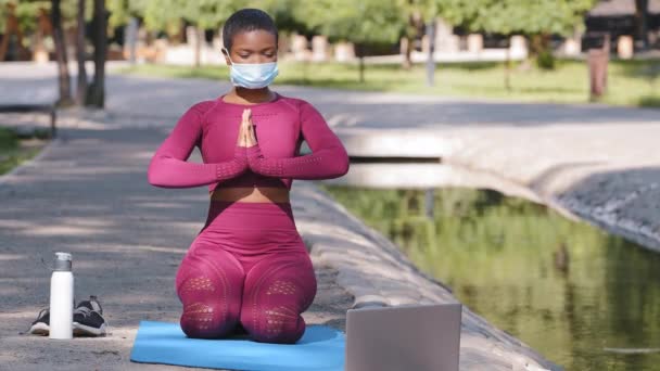 Preto focado mulher milenar sentado no tapete esporte ao ar livre usando laptop, assistindo aulas on-line. Jovem senhora em sportswear e máscara médica protetora exercitar olhando fitness vídeo na Internet — Vídeo de Stock