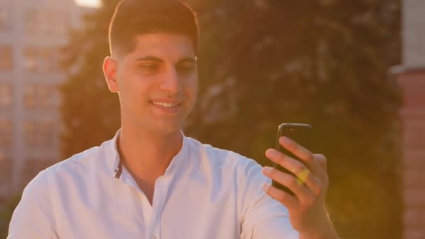 Smiling millennial Indian Arabic male student holding phone, making video call, talking to parents, girlfriend or wife, looking at camera outdoors. Young adult guy sends air kiss goodbye. Backlight — Stock Video