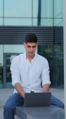 Genç, çekici Hintli Arap adam dışarıda oturuyor, dizüstü bilgisayar kullanıyor, ekrana bakıyor, e-postaları kontrol ediyor, sosyal ağdaki arkadaşlarıyla mesajlaşıyor, rapor ya da testlere hazırlanıyor, çevrimiçi, dikey video eğitimi alıyor.