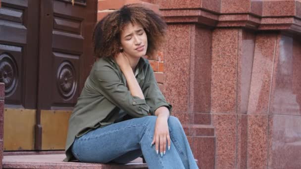 Mujer afroamericana cansada joven se sienta en las escaleras masajeando el cuello frotando los músculos tensos después de un largo viaje a pie por la ciudad. Postura incorrecta, estilo de vida sedentario, necesidad de ejercicios o concepto de tratamiento — Vídeo de stock